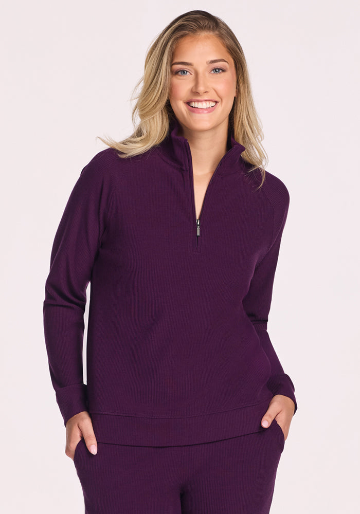 A blonde woman smiles with her hands in her pockets, wearing the Woolx Quinn Ribbed 1/4 Zip in Majestic Purple against a light background. 
