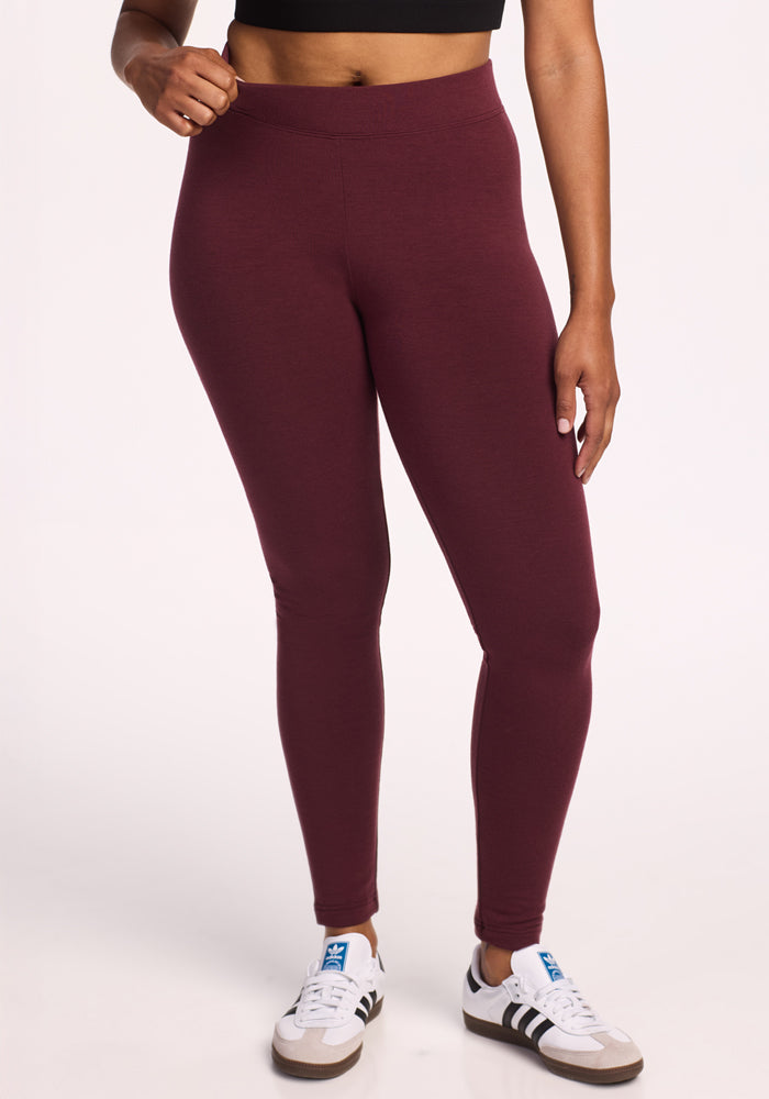 A person wearing a black crop top, Woolx Stella Leggings in Toasted Garnet, and white sneakers with black stripes stands against a plain light background. Only the body from shoulders down is shown.