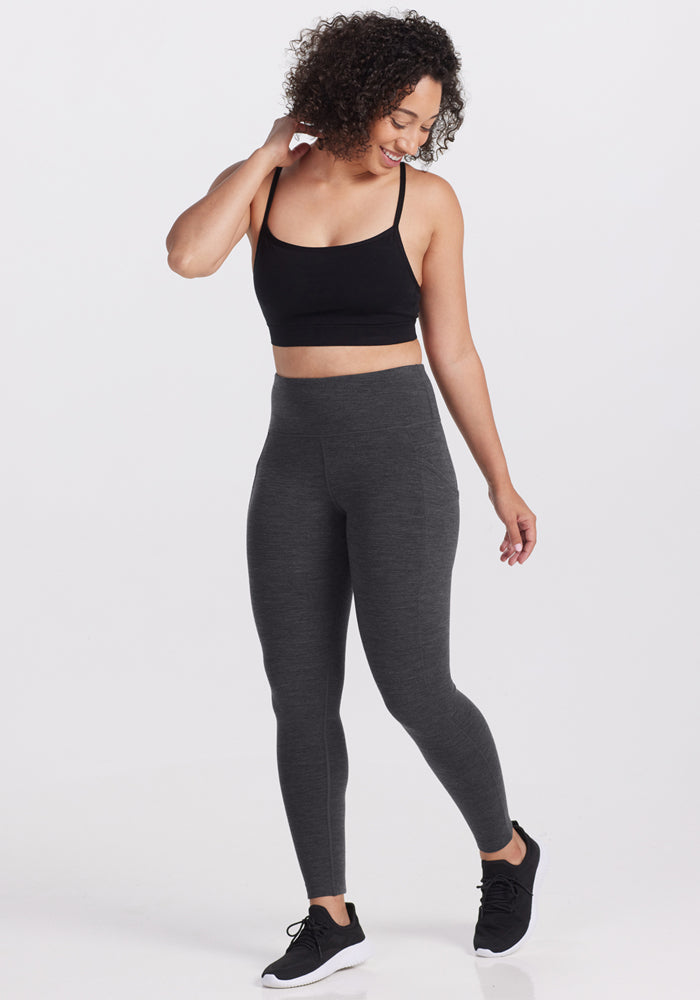 A woman with curly hair stands against a white background, smiling and touching her hair while wearing Woolx Piper Pocket Petite - Cinder Melange leggings, a black sports bra, and black sneakers.