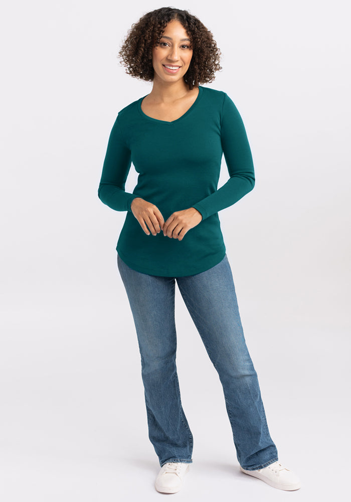 A woman with curly hair smiles, standing forward in a Woolx Eva Tunic - Riverbank Green, blue jeans, and white sneakers against a plain light background.