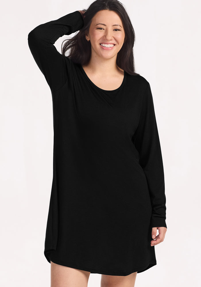 A woman with long dark hair smiles while wearing the Woolx Lucille Long Sleeve Nightgown in black, standing against a light background and gently touching her hair with one hand.