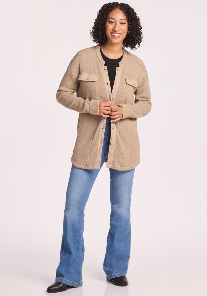 A smiling woman with curly hair wears the Woolx Oaklynn Waffle Shacket in Biscotti over a black shirt, blue flared jeans, and black shoes, standing against a plain white background.