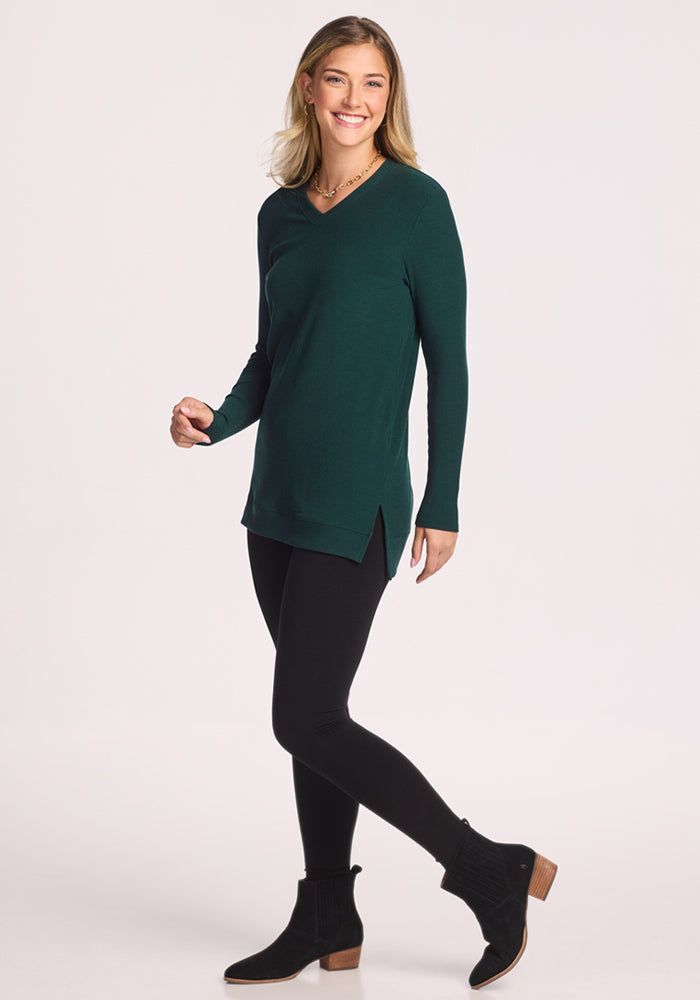 A woman smiles, wearing the Woolx Evie Ribbed Tunic in Deep Timber—a dark green long-sleeve V-neck—paired with black leggings and low-heel ankle boots, standing against a plain white background.
