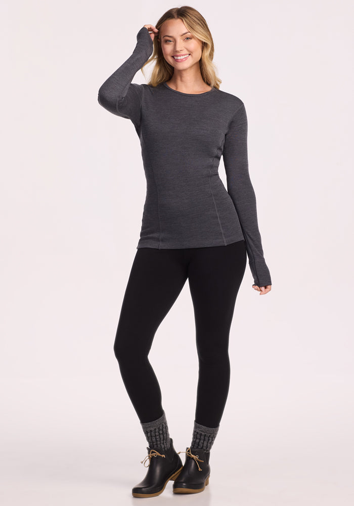 A woman stands smiling in a Woolx Hannah Long Sleeve Top in Cinder Melange, styled with black leggings, gray knit socks, and black ankle boots with tan soles, against a plain light background.