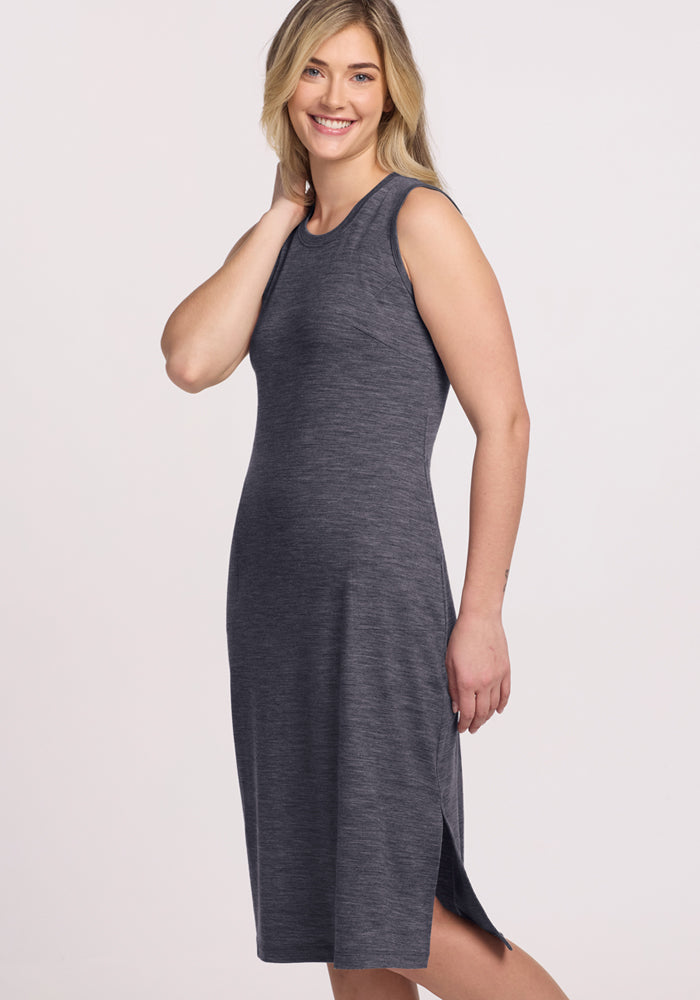 A woman with long blonde hair smiles, standing sideways and touching her neck. She wears the Woolx Cassie Dress in Cinder Melange—a sleeveless, knee-length Merino wool dress with a side slit—ideal for wrinkle-resistant travel.