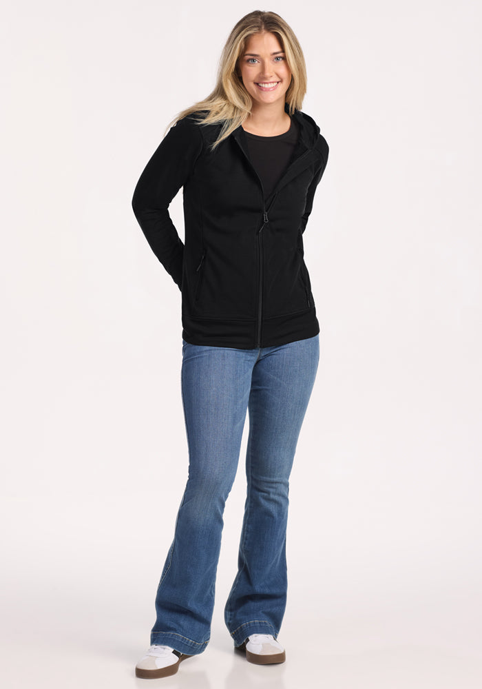 A smiling woman with long blonde hair wears the Woolx Darby Zip Up Hoodie in black, styled with blue jeans and white sneakers. She stands against a plain white background with her hands behind her back.