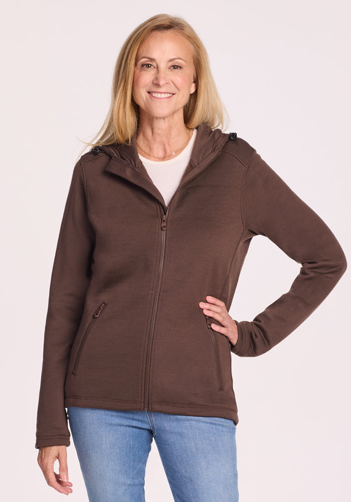A smiling woman with long blonde hair wears the Woolx Cubby Hooded Sweatshirt in Bracken over a light pink top and blue jeans, standing with one hand on her hip against a plain light background.