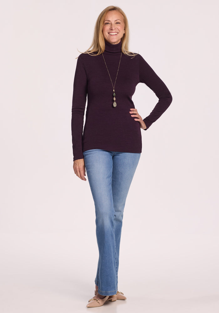 A woman with long blonde hair wears the Woolx Peyton Turtleneck in Deep Plum, light blue jeans, beige flats, and a pendant necklace, smiling with one hand on her hip against a light background.