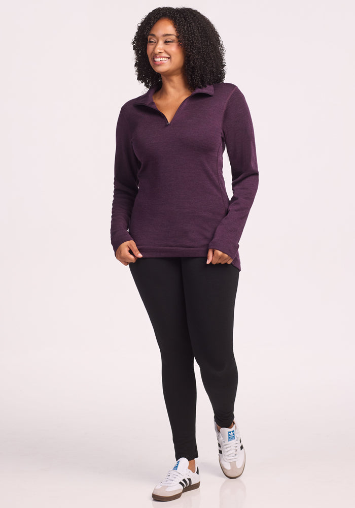 A woman with curly hair smiles while standing in front of a plain white background. She is wearing the Woolx Brooke 1/4 Zip in Deep Plum, paired with black leggings and white sneakers featuring black stripes.