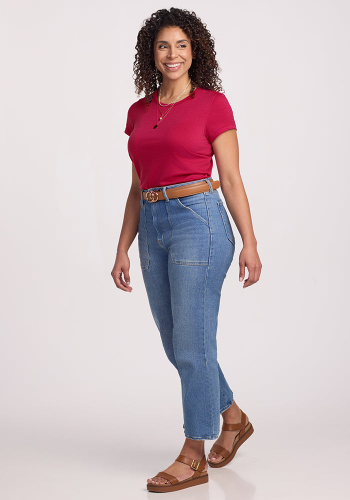 A woman with curly hair, smiling in a Woolx Addie Crew Neck - Poppy Spark, blue high-waisted jeans, brown belt, and tan sandals stands against a plain white background.