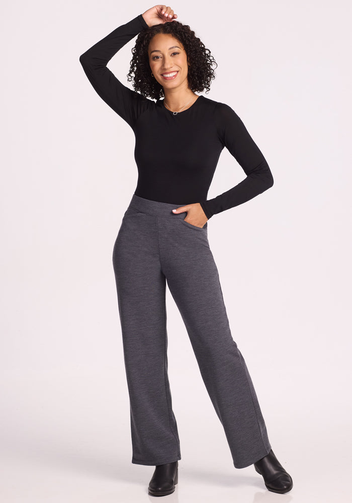 A woman with curly hair, wearing a black long-sleeve top and Woolx Ellie Wide Leg Pants in Cinder Melange, stands smiling with one hand on her head and the other in her pocket against a plain white background.