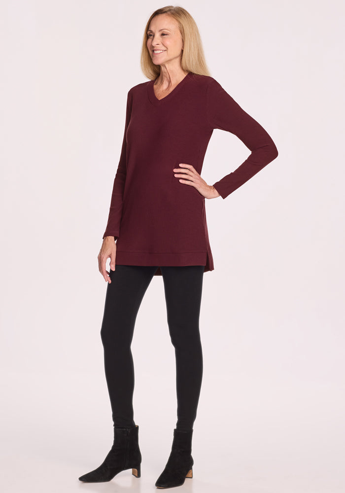 A woman with long blonde hair wears the Woolx Evie Ribbed Tunic in Toasted Garnet, paired with black leggings and ankle boots. She stands sideways, smiling slightly against a plain light background.