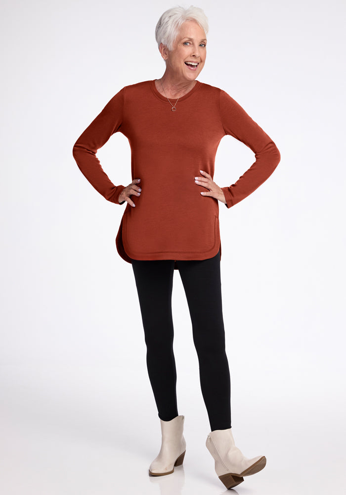 Smiling older woman with short white hair wears the Woolx Blair Tunic in Maple Leaf, black leggings, and white ankle boots, standing hands on hips against a plain white background.
