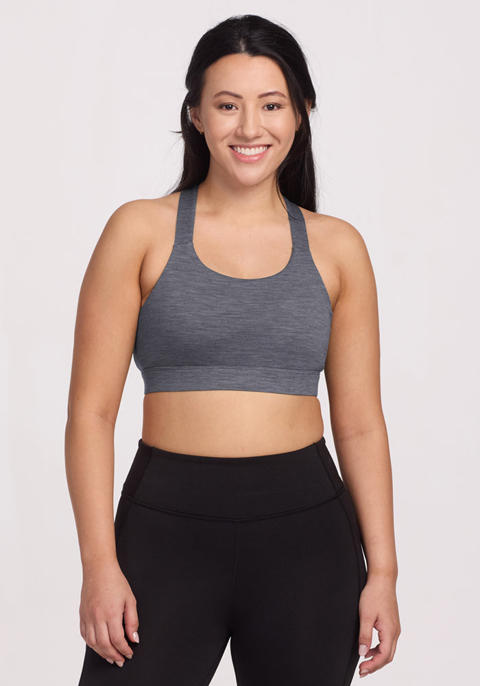 A woman with long dark hair smiles while wearing the Woolx Livi Bra in Graphite Heather and black high-waisted leggings, standing against a plain light background.