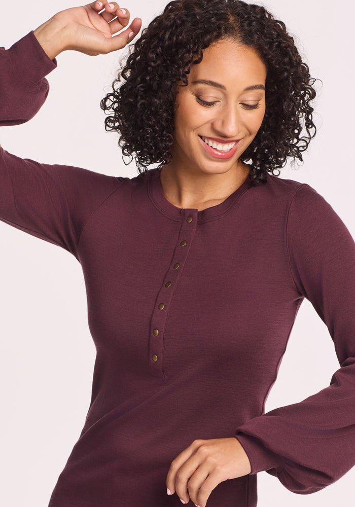 A woman with curly dark hair smiles and raises one arm while wearing the Woolx Charlotte Puff Sleeve Top in Chocolate Fig, featuring Henley-style buttons, against a plain light background.