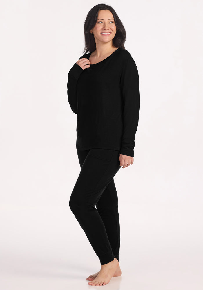 A woman with long dark hair smiles barefoot in front of a white background, wearing the Woolx Ashlie Lounge Top in black with matching pants. The wrinkle-resistant top features long sleeves for a polished, comfortable look.