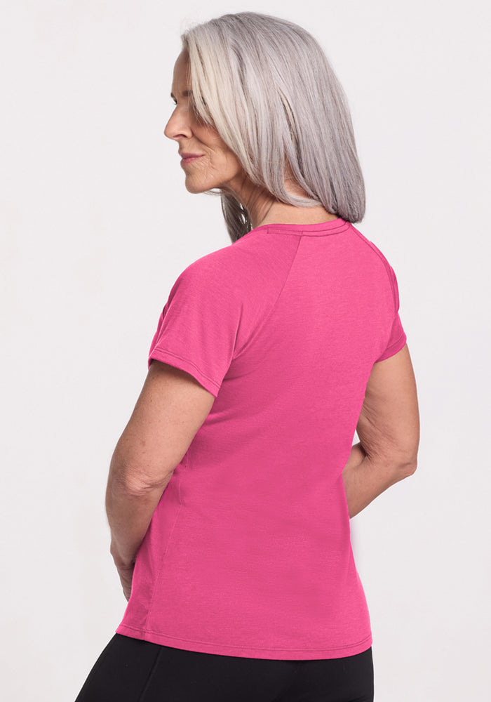 A woman with long gray hair wears the Woolx Mia V Neck in Raspberry Fizz and black pants, standing slightly turned away from the camera against a plain white background.