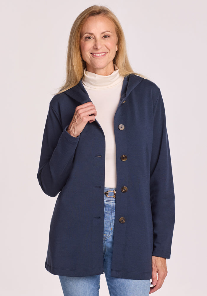 A woman with long blonde hair smiles while wearing the Woolx Edna Hooded Cardigan in Deep Navy over a white turtleneck and light blue jeans, standing against a plain background—ideal for chilly days. 