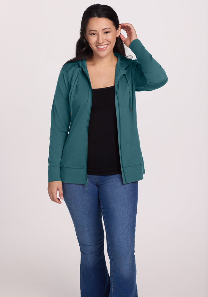 A woman with long dark hair smiles, wearing the Woolx Ryann Hoodie in Tidepool over a black top and blue jeans—an essential travel piece—while standing against a plain light background with one hand touching her hair.