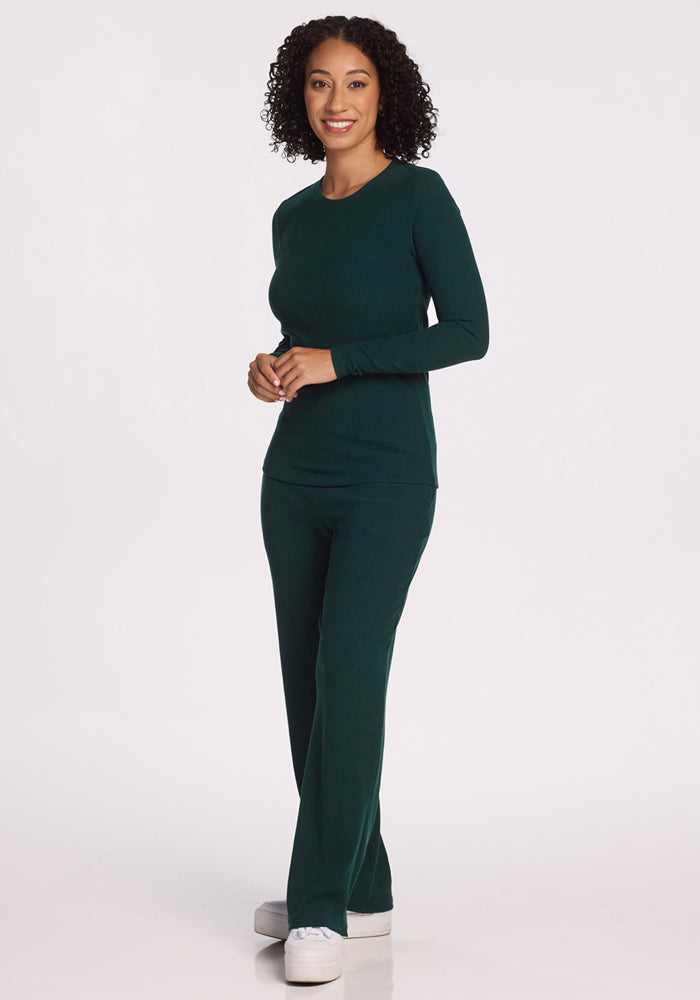 A woman with curly hair smiles while wearing the Woolx Hadley Ribbed Crew in Deep Timber and matching pants, paired with white sneakers, against a plain white background.