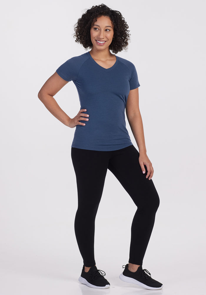 A woman with curly hair stands smiling, wearing the Woolx Mia V Neck - Starry Night moisture-wicking tee, black leggings, and black sneakers against a plain white background.