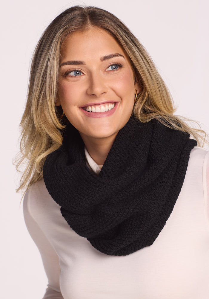 A smiling woman with blonde hair wears the Woolx Noelle Infinity Scarf in black over a light long-sleeve top, highlighting this cozy winter accessory against a plain background.