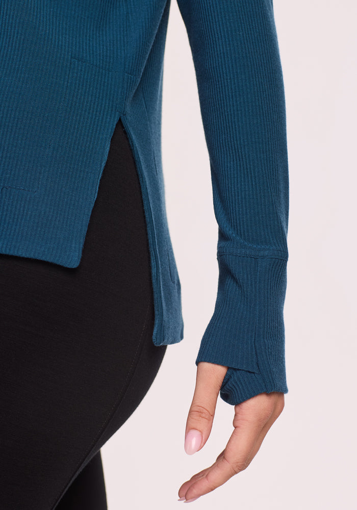 A close-up of a person wearing the Woolx Ainsley Sweater in Majolica Blue with thumbholes, ribbed texture, and side slit, paired with black leggings. Their hand is relaxed, displaying neatly manicured nails.