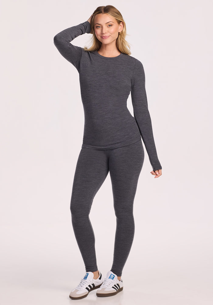 A woman smiles in the Woolx Sadie Baselayer Top in Cinder Melange, paired with matching leggings and white sneakers. She touches her head and poses against a simple, light background.