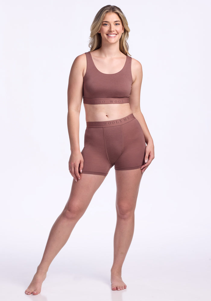 A woman with long blonde hair smiles while standing barefoot in a matching mauve sports bra and Woolx Elliott Boyshort - Nutmeg set, against a plain white background.
