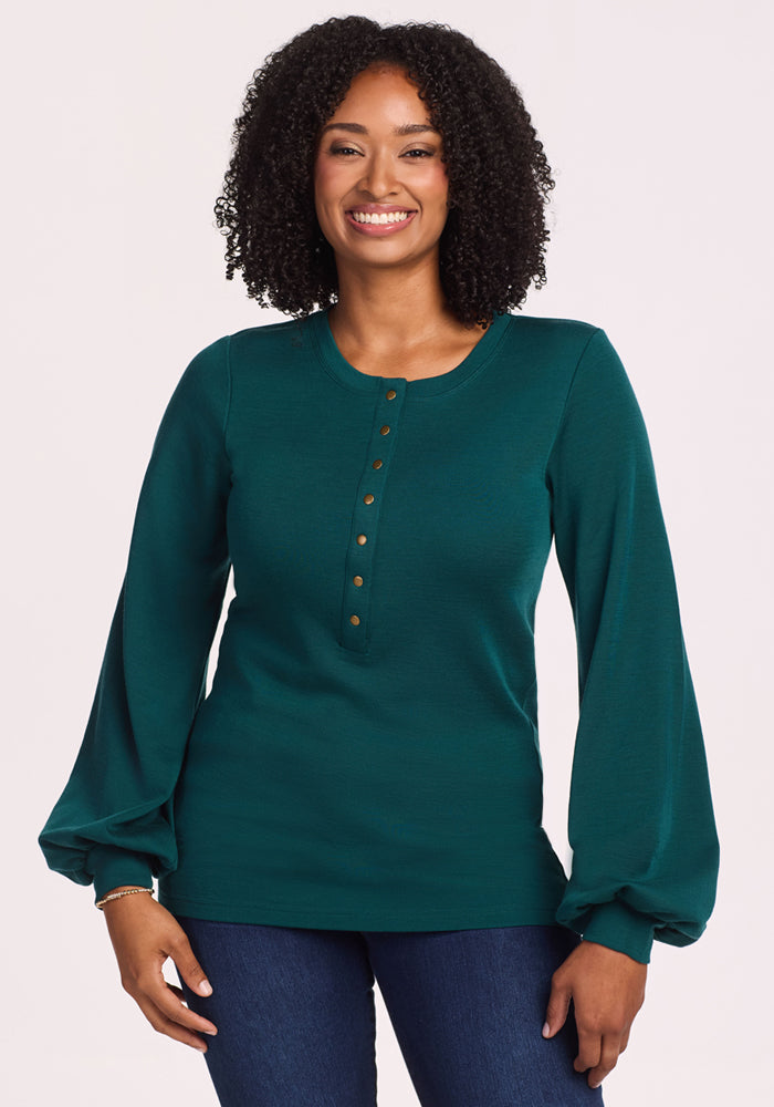 A woman with curly hair smiles while wearing the Woolx Charlotte Puff Sleeve Top in Riverbank Green with Henley buttons and balloon sleeves, paired with blue jeans, standing against a plain light background. 