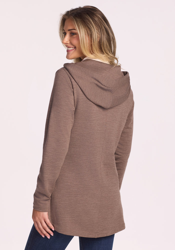 A blonde woman smiles over her shoulder, wearing the Woolx Edna Hooded Cardigan in Simply Taupe with dark jeans, posed against a plain light background.