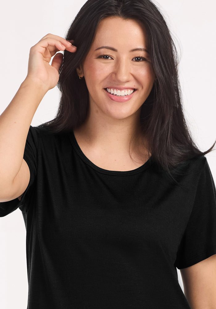 A woman with long dark hair, wearing the Woolx Desi Nightgown in black, smiles at the camera and touches her hair with one hand against a plain light background.