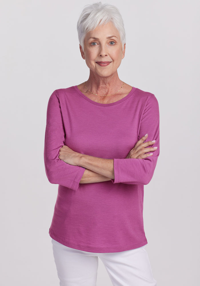 An older woman with short white hair and a confident smile stands with arms crossed, wearing the Woolx Jenny 3/4 Sleeve Crew Neck in Purple Wine and white pants against a light gray background. 