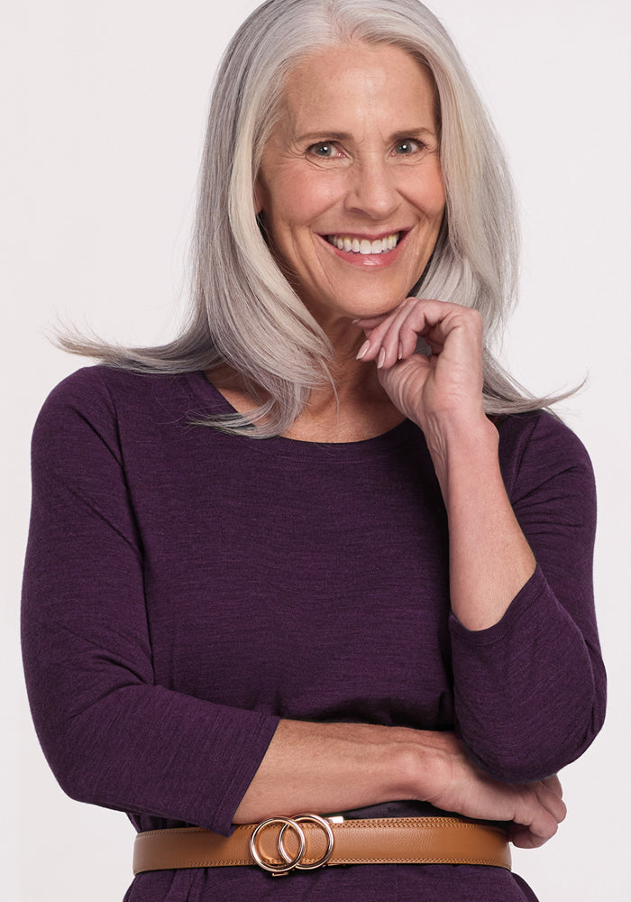 A smiling older woman with long gray hair models the Woolx Rissa A Line Dress in Deep Plum, a machine washable Merino wool style ideal for fall and winter layering, paired with a brown belt against a plain light background.