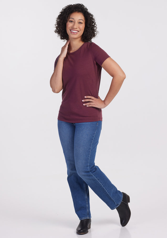 A woman with curly hair smiles, wearing the Woolx Liza Crew Neck Tee in Grape Harvest, paired with blue jeans and black boots. She stands on a plain white background, one hand on her hip and the other touching her neck.