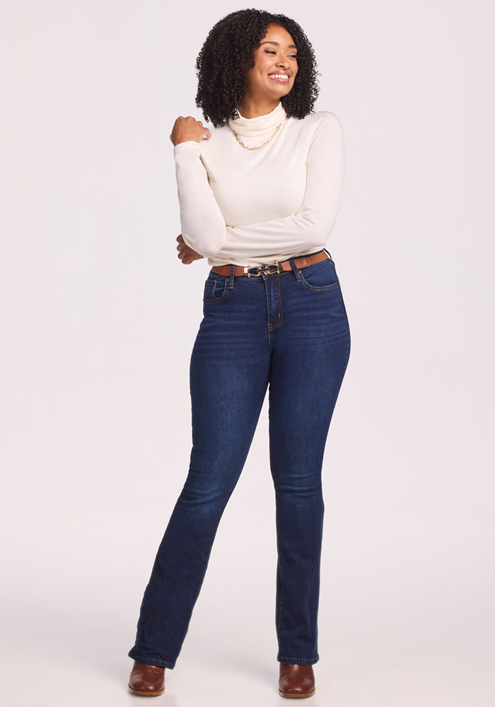 A woman with curly hair wears the Woolx Kennedy Bodysuit in Buttercream, paired with dark blue jeans, a brown belt, and brown boots. She stands smiling with one hand on her hip against a plain white background.