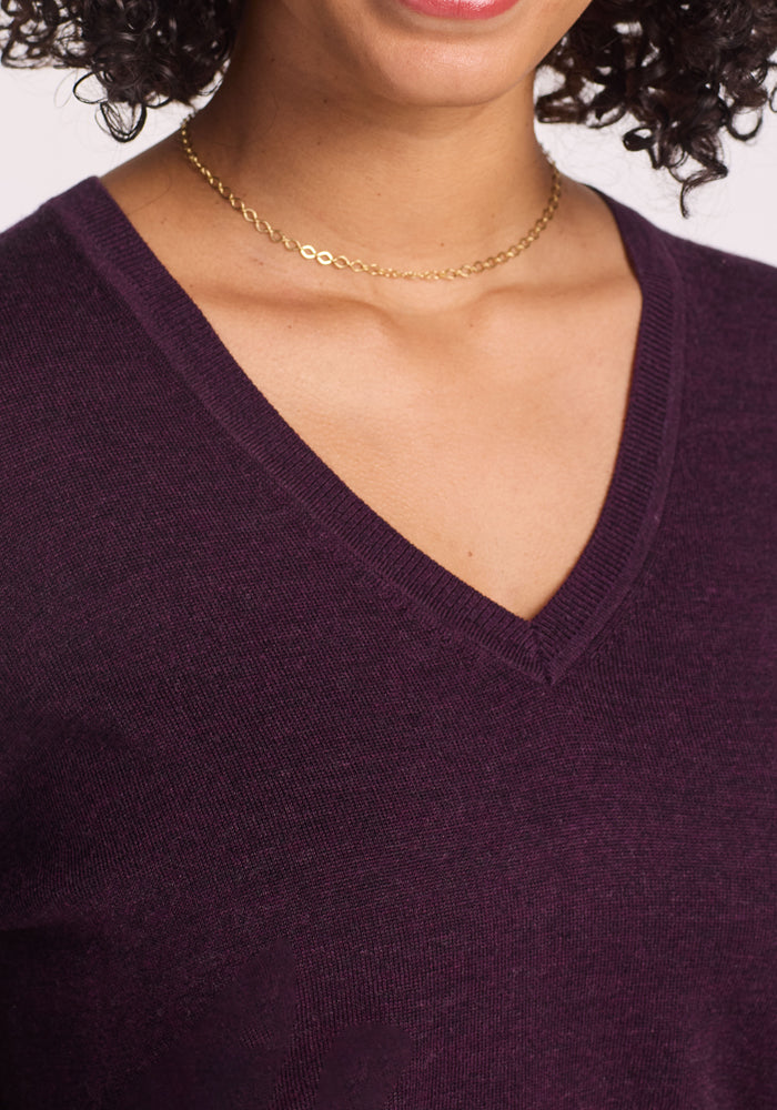 A woman wears the Woolx Francesca Sweater in Deep Plum, featuring a V-neck Merino wool design, along with a delicate gold chain necklace. Only her neck, lower face, and upper chest are visible, with curly dark hair.