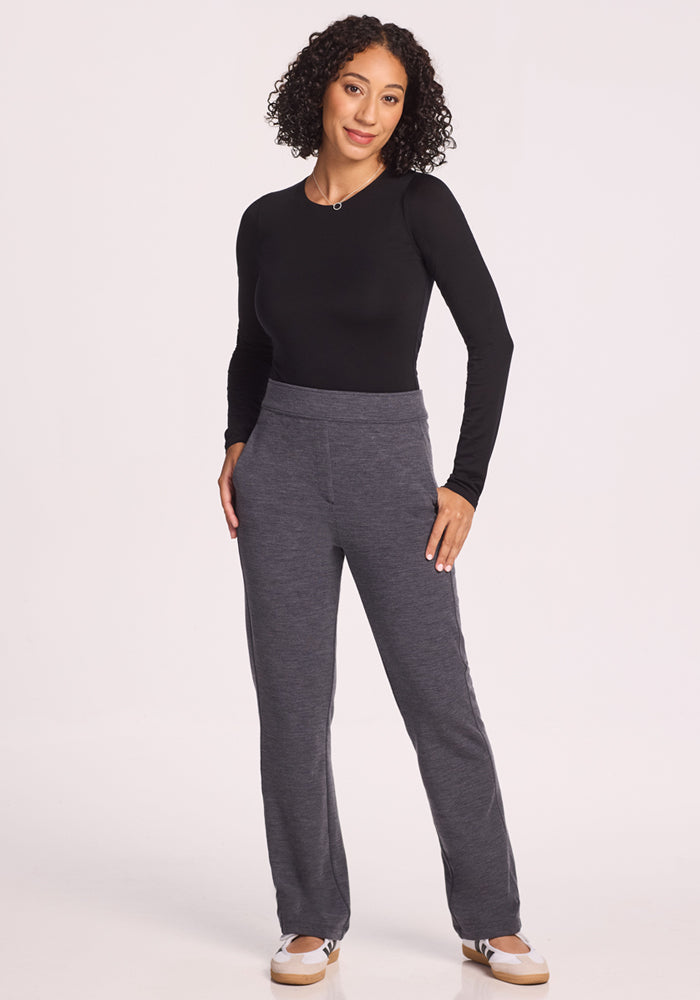 A woman with curly hair smiles, wearing a fitted black long-sleeve top, Woolx Emerson Straight Leg Pants in Cinder Melange, and white sneakers against a plain light background.
