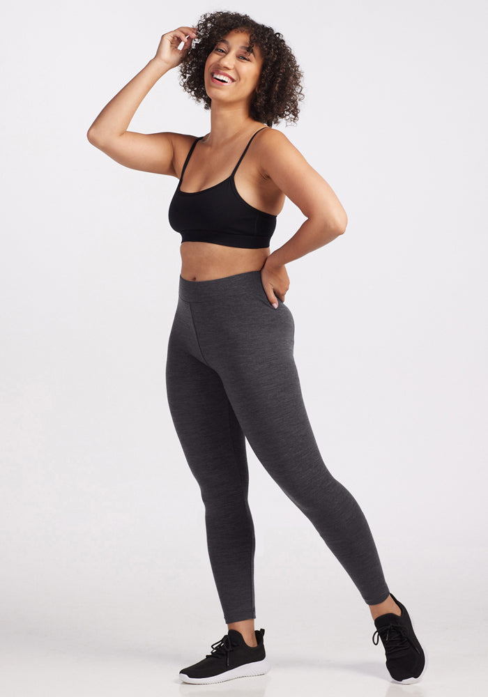 A smiling woman with curly hair stands against a plain background, wearing Woolx Stella Petite - Cinder Melange leggings, a black sports bra, and black sneakers. One hand is on her hip while the other touches her hair.