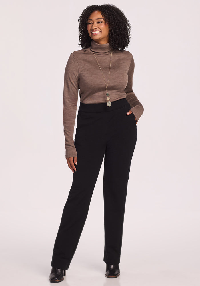A woman with curly hair wears the Woolx Peyton Turtleneck in Simply Taupe, paired with black high-waisted pants, ankle boots, and a long pendant necklace. She smiles with one hand in her pocket against a plain light background.