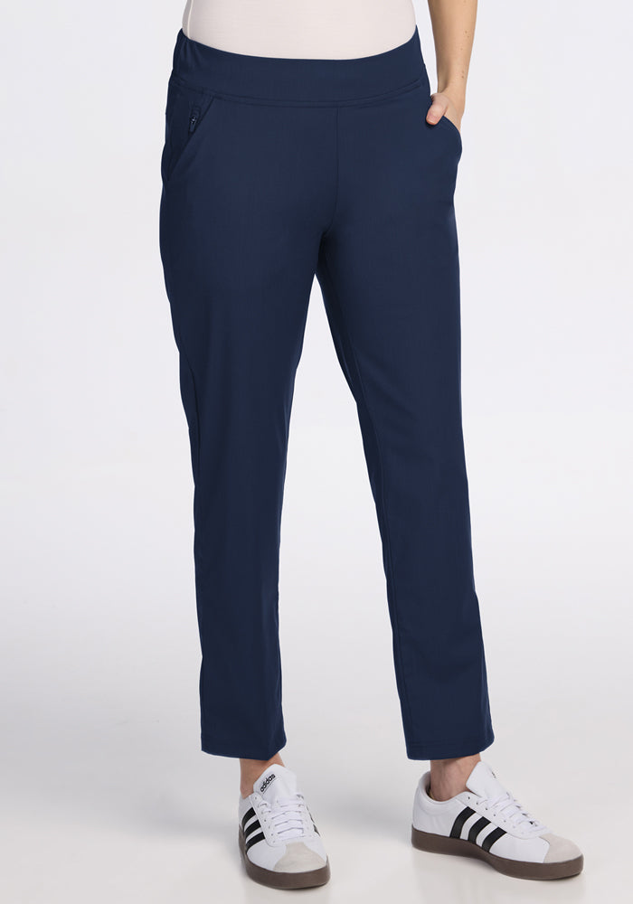 A person wears Woolx Nicole Ankle Pants in Deep Navy, paired with white sneakers featuring black stripes, standing against a plain white background. 