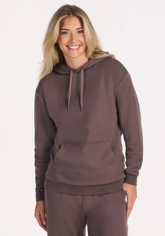 A person with long blonde hair smiles while wearing the Woolx Avery Hoodie in Plum Truffle, standing against a plain light background. 