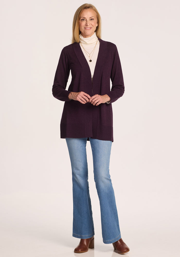 A woman smiles in the Woolx Macie Sweater in Deep Plum, layered over a white turtleneck with blue jeans and brown ankle boots, accessorized with layered necklaces against a plain white background.