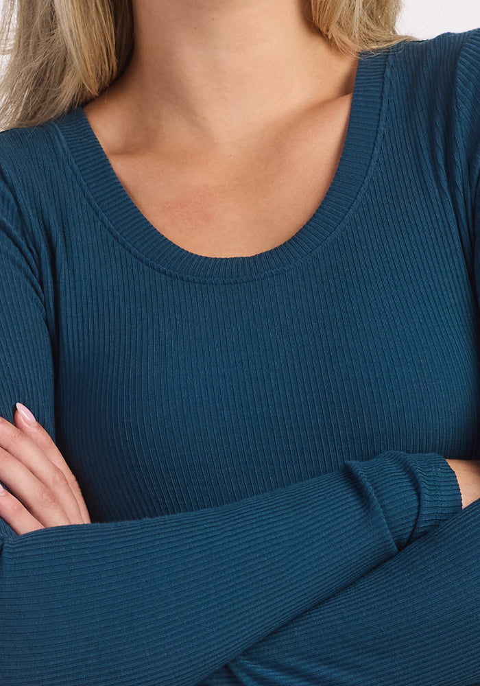 A person with long blonde hair stands with their arms crossed, wearing the Woolx Caroline Ribbed Twirl Dress in Majolica Blue. Their face is not visible in the image.