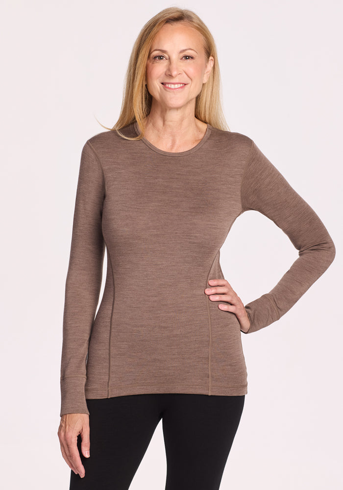 A smiling woman with long blonde hair models the Woolx Hannah Long Sleeve Top in Simply Taupe and black pants, posing with one hand on her hip against a plain light background. 