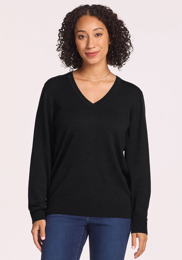 A woman with curly hair wears the Woolx Francesca Sweater in black and blue jeans. Against a plain light background, she smiles slightly at the camera, effortlessly showcasing this travel-friendly sweater. 