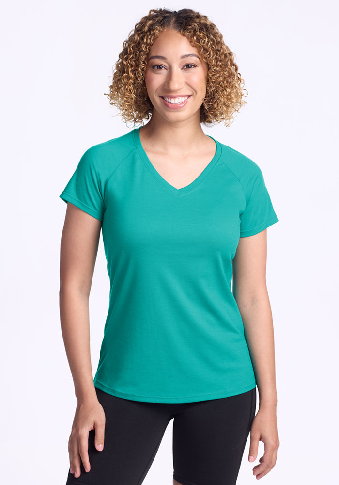 A smiling woman with curly hair wears the Woolx Mia V Neck - Cool Jade, a women’s moisture-wicking Merino wool t-shirt, paired with black pants against a plain white background. 