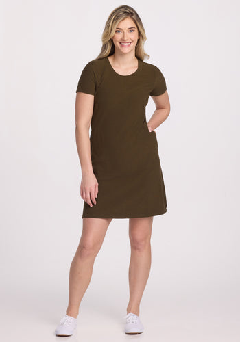 A person stands against a plain background, clad in the Harlowe Ribbed Dress - Olive Leaf by Woolx and white sneakers. The odor-resistant merino wool dress reaches knee-length, and they smile confidently with one hand on their hip.