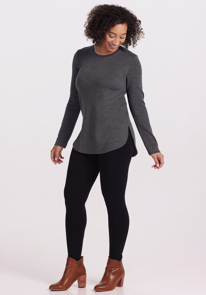 A woman stands against a plain background wearing the Woolx Blair Tunic in Cinder Melange, paired with black leggings and brown ankle boots. She looks down with a slight smile, enjoying its odor-resistant comfort and lightweight warmth.