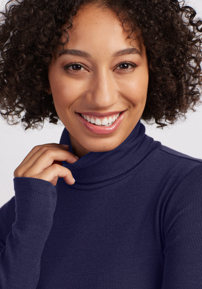 A smiling woman with curly hair wears the Woolx Sage Ribbed Turtleneck in Midnight Sapphire, touching the collar and looking at the camera against a light background.